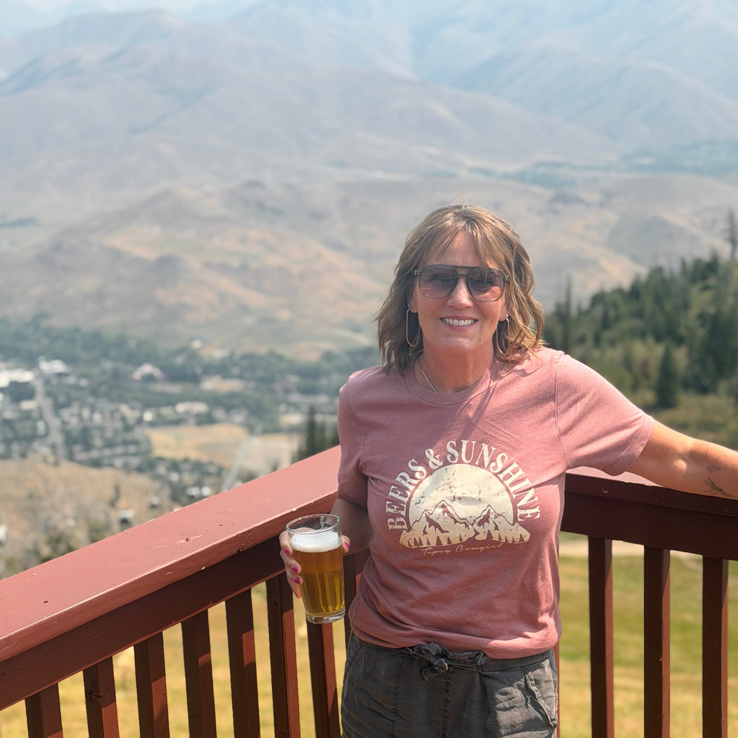 Woman wearing a mauve “Beers & Sunshine” Tipsy Cowgirl tee and gray shorts, holding a beer while standing on a mountain overlook with a scenic valley and mountain range in the background.